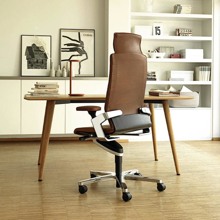 A modern home office showcasing the ON mid-century wooden desk, paired with a brown ergonomic swivel chair. Shelves lined with books and decorative accents add character, while framed artwork rests elegantly on a shelf against a crisp white wall in the background. Studio Office Cyprus
