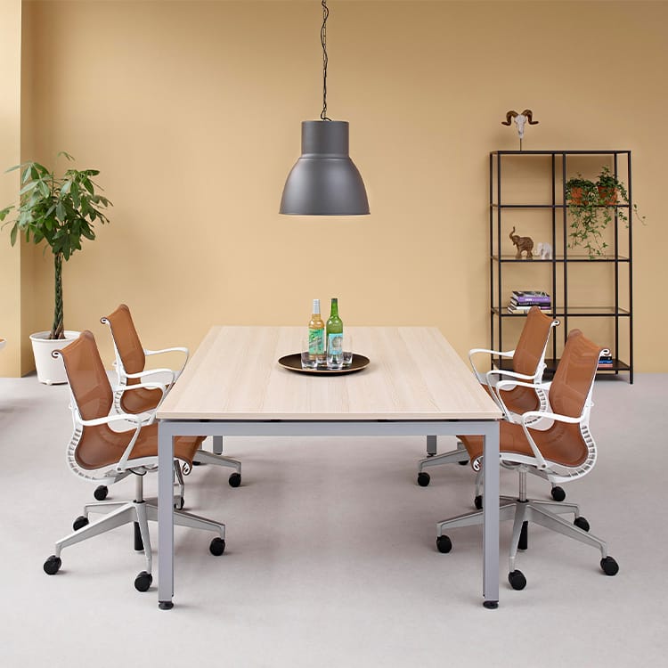 A contemporary meeting room featuring a Sense Conference Table surrounded by four brown chairs, with refreshments arranged on a tray. A pendant lamp hangs overhead, while a potted plant and a black shelf adorned with decor accent the beige wall in the background. Studio Office Cyprus