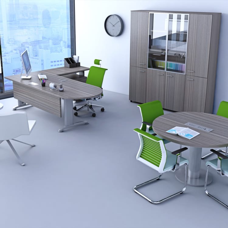 A contemporary office space bathed in natural light from expansive windows, showcasing an Ergo Managerial desk equipped with a computer, complemented by stylish green and white chairs. Nearby, a round meeting table holds neatly arranged papers and magazines. The scene is completed by a wall clock and a glass-door cabinet displaying organized folders. Studio Office Cyprus
