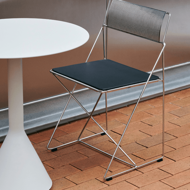 Σύγχρονη καρέκλα X-Line Chair Chrome με μαύρο κάθισμα και χρωμιωμένο σκελετό, δίπλα σε λευκό τραπέζι, σε επιφάνεια από τούβλα.