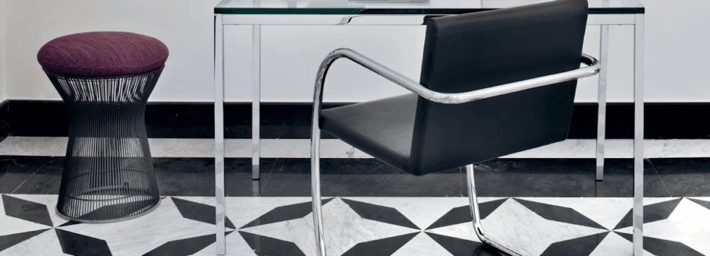 Το Platner Stool με βάση από σκούρο ατσάλινο σύρμα και ανάγλυφο μαξιλάρι σε χρώμα μούρου, δίπλα σε κομψό γραφείο με γυάλινη επιφάνεια.