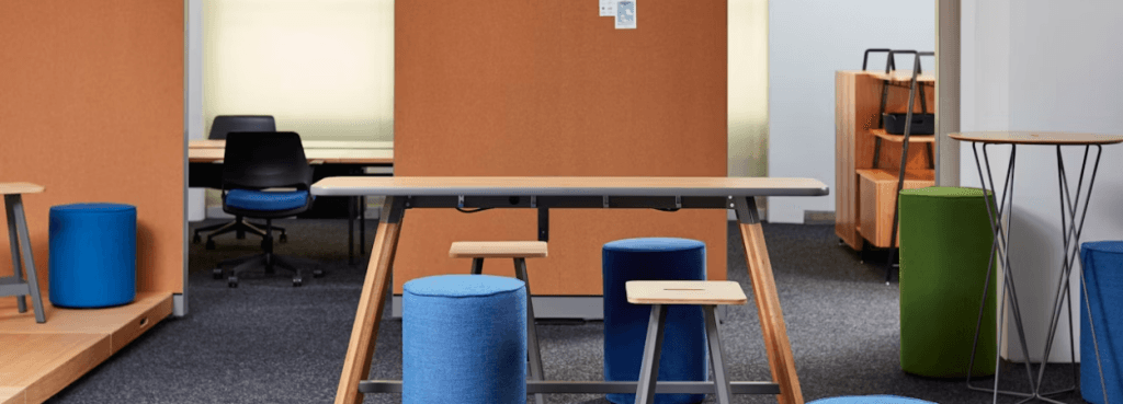 Τα Rockwell Unscripted Easy Stools σε συνδυασμό με πολύχρωμους υφασμάτινους κυλίνδρους, γύρω από κεντρικό τραπέζι εργασίας από ξύλο και μέταλλο.
