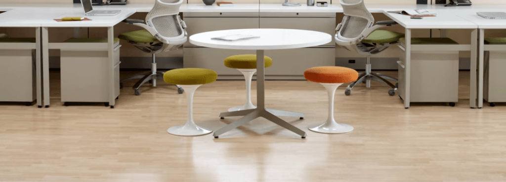 Τρία πολύχρωμα σκαμπό Knoll Tulip Stool με υφασμάτινα μαξιλάρια γύρω από λευκό τραπέζι σε φωτεινό χώρο εργασίας.